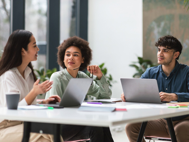 Three young colleagues of mixed ethnicity sitting at table with laptops in a modern office, collaborating on a project. Coworkers enjoying cooperative professional team work