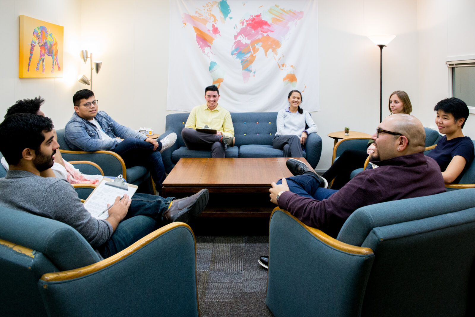 A group of people sitting in a small room with comfy chairs having a conversation.
